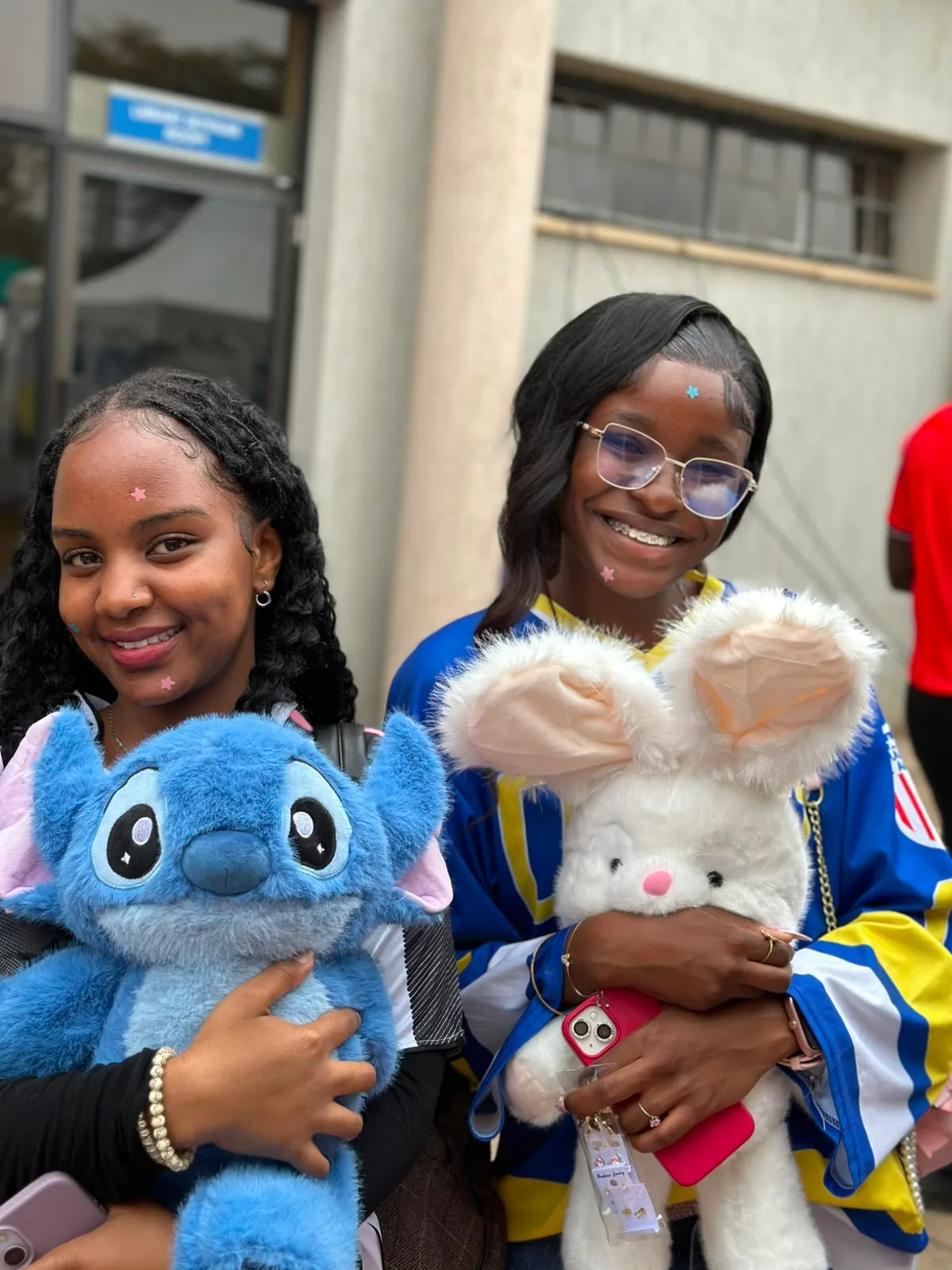 Two friends smiling with matching star stickers, holding a Stitch plushie and a white bunny plushie