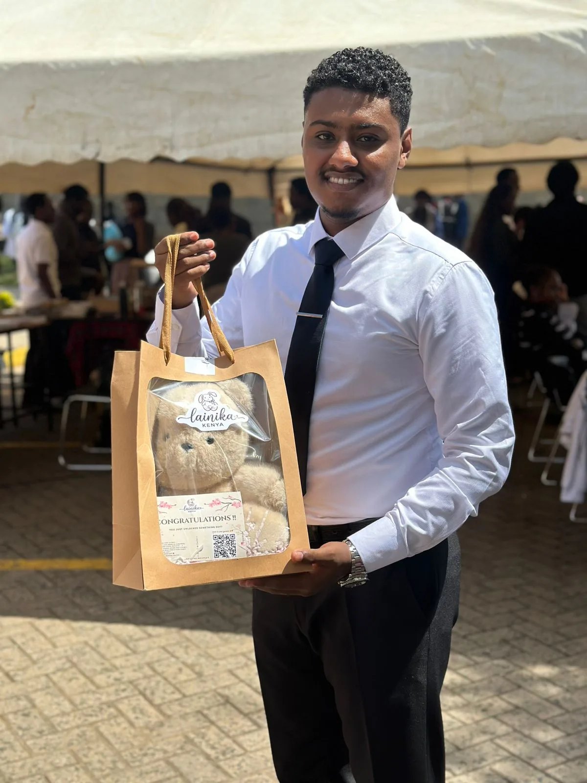 A young man in a shirt and tie holding a Lainika gift bag with a teddy bear visible inside
