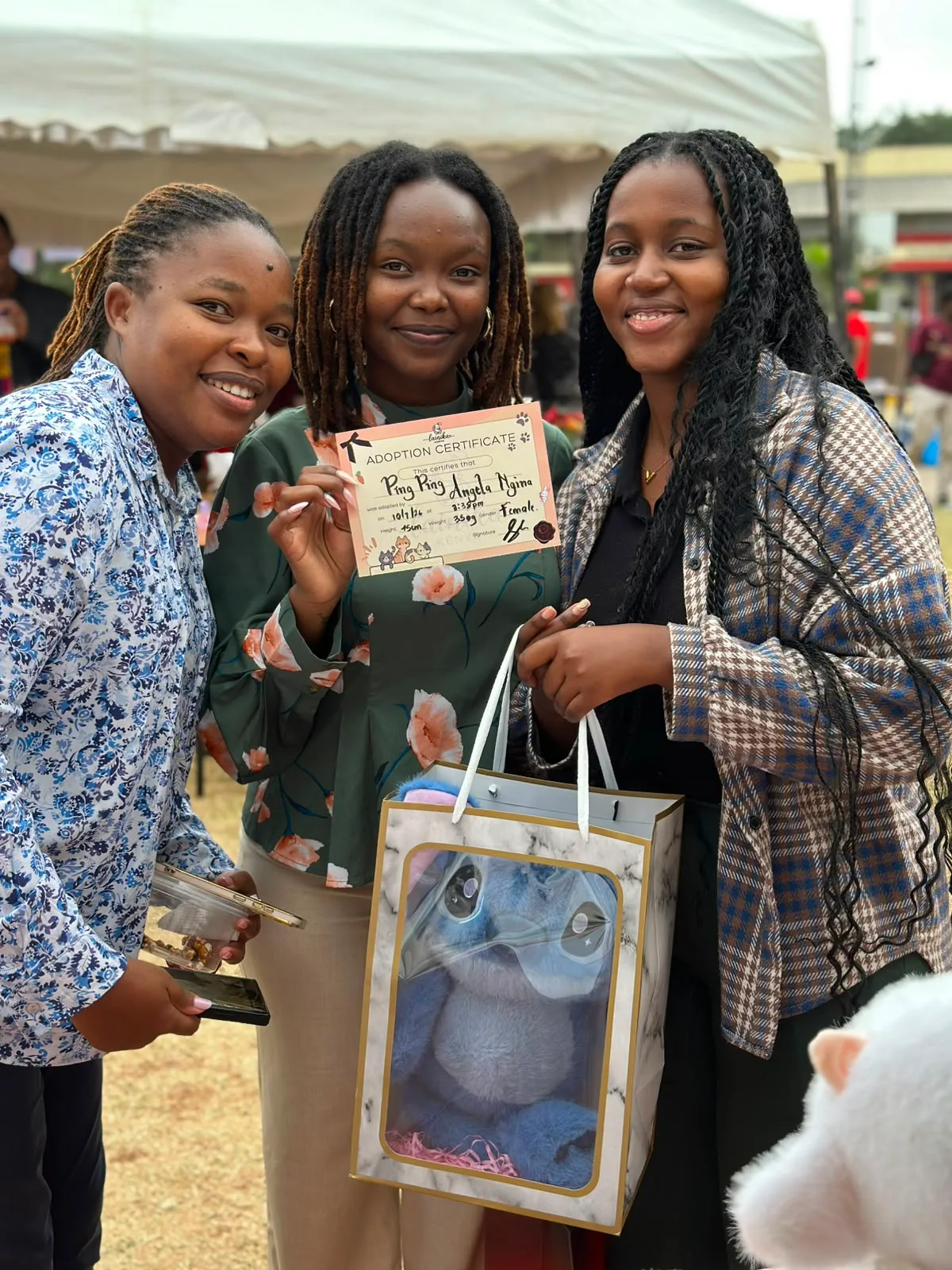 Three friends smiling, holding a Lainika Signature Wrap gift bag and adoption certificate