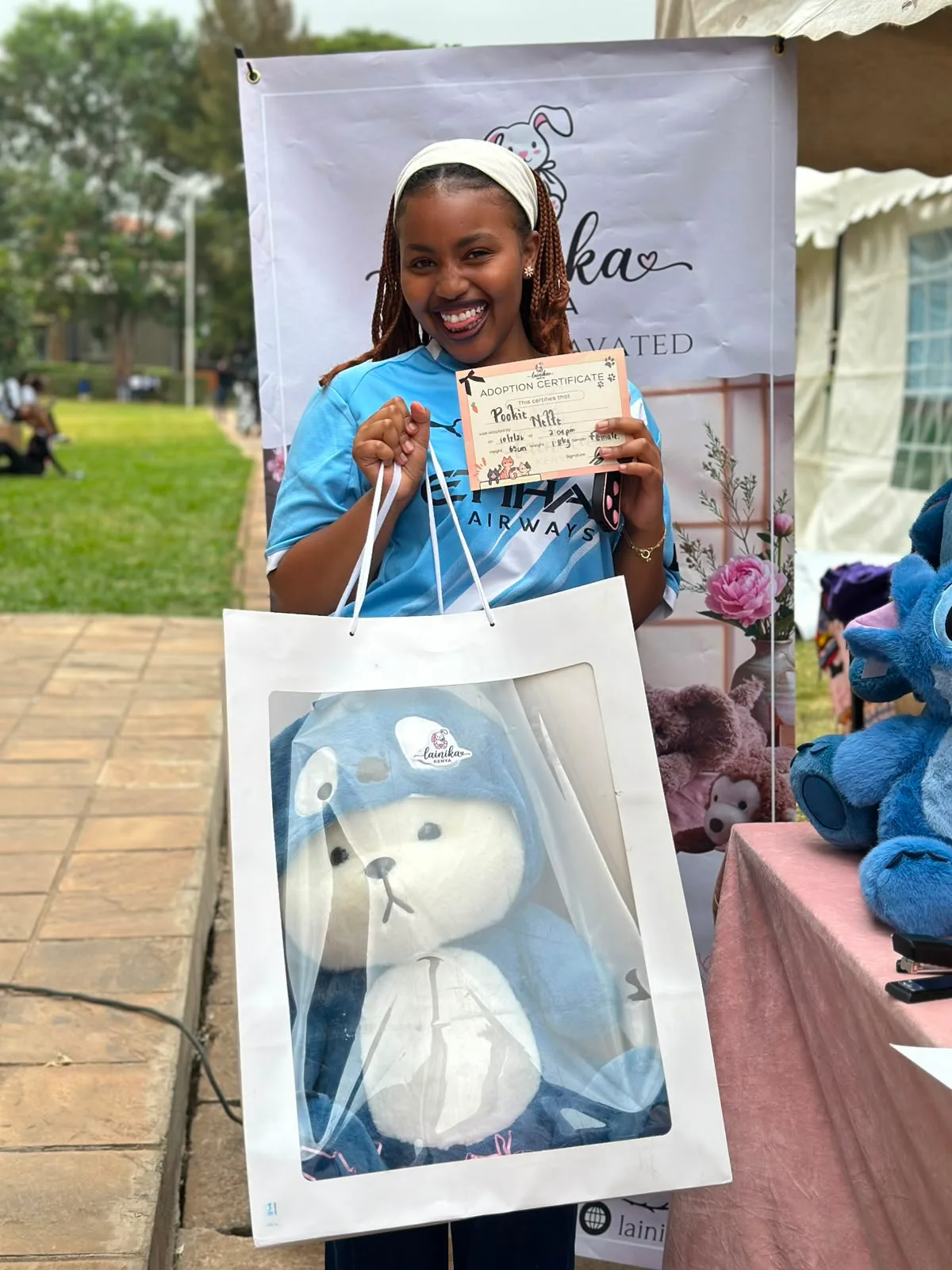 A woman smiling at the Lainika stall, holding an adoption certificate for her XL Stitch plushie named Pookie