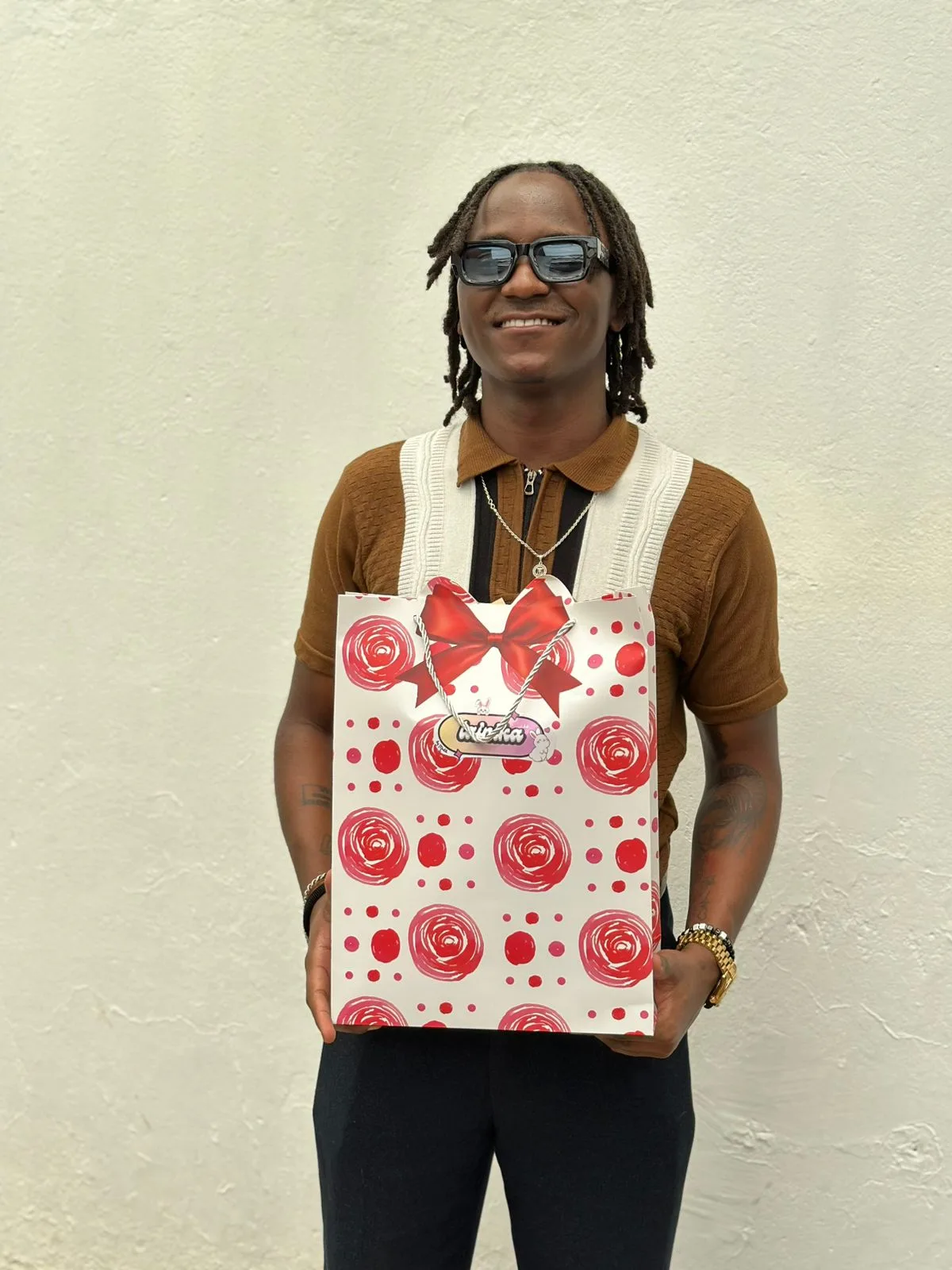 A man in sunglasses and dreadlocks smiling while holding a rose-print Lainika gift bag with a red bow